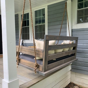 May include: A wooden swing bench with a gray stain hangs from a porch ceiling with thick rope. The swing has a gray cushion and several white throw pillows. The porch is made of wood and has a white railing.