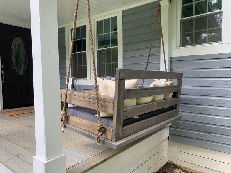 May include: A wooden swing bench with a gray stain hangs from a porch ceiling with thick rope. The swing has a gray cushion and several white throw pillows. The porch is made of wood and has a white railing.