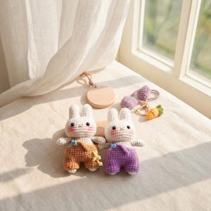 May include: Two handmade crochet bunny dolls with white bodies, rosy cheeks, and stitched facial features. One bunny wears orange overalls, the other purple. A wooden disc, a purple keychain, and a carrot keychain are also visible.