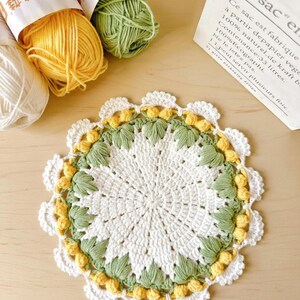 May include: A handmade, white crochet doily with a scalloped edge. The doily features a central design with green leaves and yellow accents. Yarn skeins in yellow, green, and white are visible in the background.