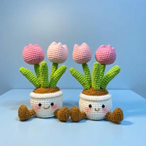 May include: Two crochet tulip plants with pink and light pink flowers, green leaves, and smiling white pots. The pots have brown soil and brown feet. The background is a light blue.
