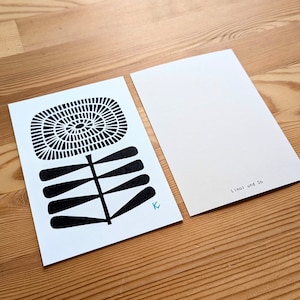 May include: Two white postcards on a wooden surface. One postcard features a black and white floral design with a spiral center and a stem with leaf-like shapes. The other postcard is blank with the text "Linol und So". A small, light blue "K" is visible.