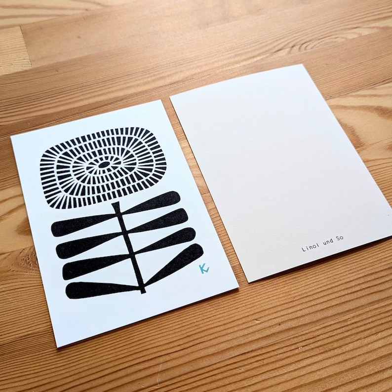 May include: Two white postcards on a wooden surface. One postcard features a black and white floral design with a spiral center and a stem with leaf-like shapes. The other postcard is blank with the text "Linol und So". A small, light blue "K" is visible.