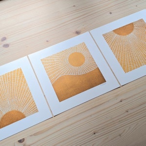 Könnte beinhalten: Drei gerahmte Kunstdrucke mit einem Sonnenmotiv in Gold und Weiß. Die Sonne wird mit strahlenförmigen Linien und einem kreisförmigen Zentrum dargestellt. Die Drucke sind auf weißem Papier und werden auf einer hellen Holzoberfläche präsentiert.