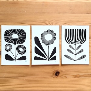 May include: Three black and white floral art prints on white paper. Each print features a different stylized flower design. The prints are displayed on a wooden surface.
