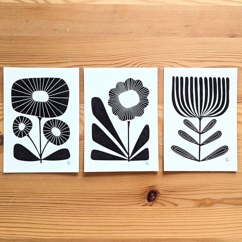 May include: Three black and white floral art prints on white paper. Each print features a different stylized flower design. The prints are displayed on a wooden surface.
