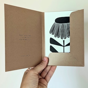 May include: A greeting card with a black and white floral design. The card is inside a brown paper envelope. The text on the card reads "K&uuml;ss' die Erde, in der Du die Blume bist." and "Linol und So."