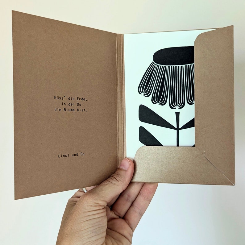 May include: A greeting card with a black and white floral design. The card is inside a brown paper envelope. The text on the card reads "K&uuml;ss' die Erde, in der Du die Blume bist." and "Linol und So."