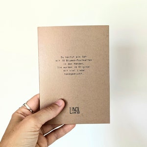 May include: A set of 10 flower postcards in a person's hand. The postcards are printed on brown cardstock. The text on the card reads: "You are holding a set with 10 flower postcards in your hands. They were originally hand-printed with much love."