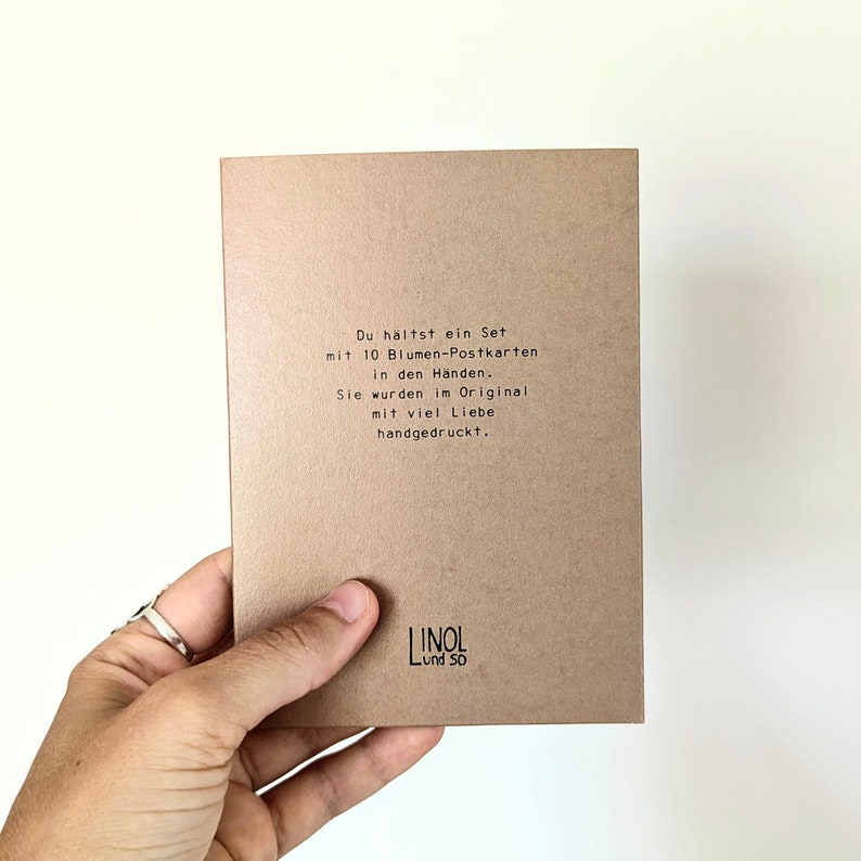 May include: A set of 10 flower postcards in a person's hand. The postcards are printed on brown cardstock. The text on the card reads: "You are holding a set with 10 flower postcards in your hands. They were originally hand-printed with much love."