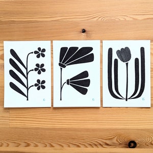 May include: Three minimalist floral art prints. Each print features a black floral design on a light blue background. The prints are displayed on a wooden surface. The floral designs include stylized flowers and leaves.