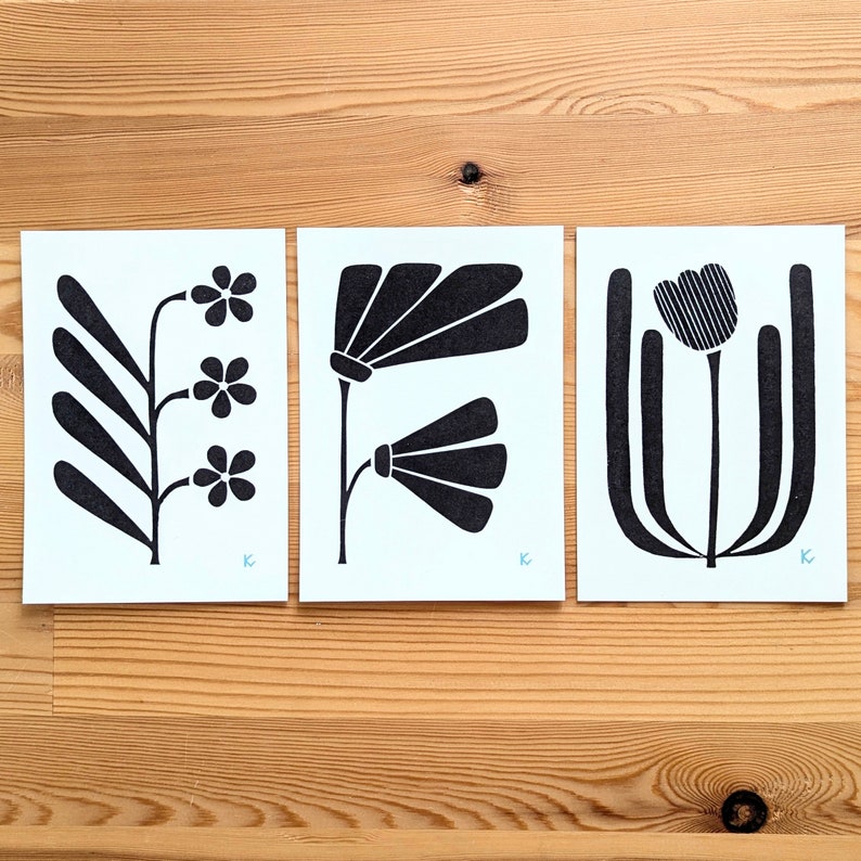 May include: Three minimalist floral art prints. Each print features a black floral design on a light blue background. The prints are displayed on a wooden surface. The floral designs include stylized flowers and leaves.