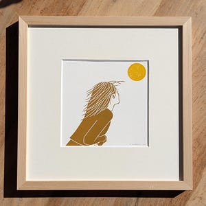 Puede incluir: Impresión artística enmarcada que presenta una ilustración estilizada de una persona mirando hacia un sol amarillo. La figura, en tonos dorados y marrones, tiene el pelo al viento. La obra se presenta sobre fondo blanco y enmarcada en madera natural.