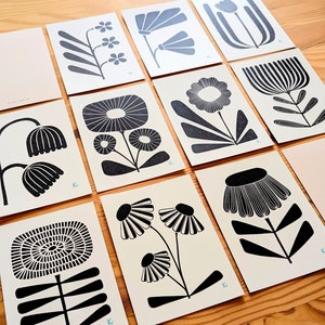 May include: A collection of floral art prints featuring various stylized black and white botanical designs. The prints are arranged on a wooden surface, showcasing a range of flower and plant illustrations. Each print has a light-colored background.