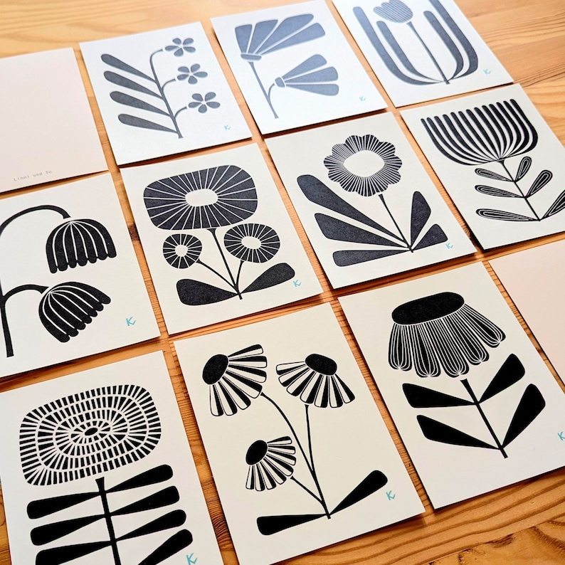 May include: A collection of floral art prints featuring various stylized black and white botanical designs. The prints are arranged on a wooden surface, showcasing a range of flower and plant illustrations. Each print has a light-colored background.
