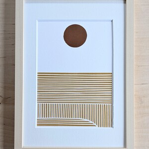 Puede incluir: Impresión artística abstracta enmarcada. La obra presenta un círculo marrón en la parte superior, con líneas doradas horizontales y una sección inferior curva sobre fondo blanco. El marco es de madera clara natural.