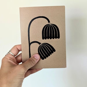May include: A tan-colored card with a black floral design. The design features two stylized flower heads connected by a curved stem. The card is held in a hand.