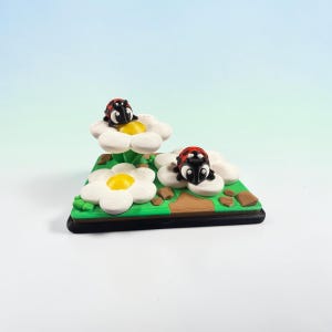 May include: A decorative figurine featuring two ladybugs with red and black spotted wings resting on white flowers with yellow centers. The flowers are set on a green base with brown accents, all mounted on a black rectangular platform.