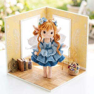 May include: A handcrafted crochet fairy doll with red hair, blue dress, and white wings. The doll is displayed in a miniature room with a gold chair, a basket of flowers, and small books. The room has yellow walls with a feather pattern.