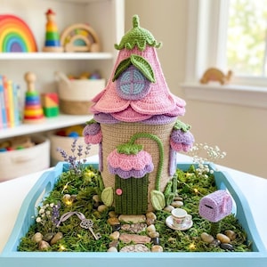 Puede incluir: Una casa de hadas tejida a ganchillo con un techo en forma de flor rosa y morada, una puerta verde y detalles florales. La casa está sobre una bandeja azul con musgo, piedras y un buzón en miniatura.