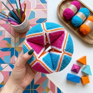 Könnte beinhalten: Ein handgefertigter, farbenfroher Häkelball in Blau, Pink und Orange. Der Ball, der in einer Hand gehalten wird, hat ein geometrisches Design. Weitere Elemente sind Häkelnadeln, Garn und geometrische Holzformen. Das Thema ist Handarbeit und Häkeln.