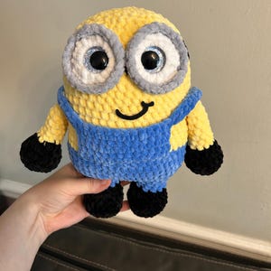 Può includere: Un giocattolo di peluche all'uncinetto che ricorda un minion del franchise Cattivissimo Me. Il giocattolo è giallo con una salopette blu, scarpe nere e grandi occhi rotondi e sporgenti.