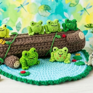May include: A collection of handmade crochet frogs in various shades of green, resting on a brown log and a blue lily pad. The scene includes small red accents and a neutral-colored frog, creating a whimsical, nature-themed display.