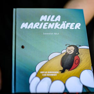 May include: A blue book with the title "Mila Marienkäfer" and the text "beweist Mut". The cover features a cartoon ladybug on a yellow flower.