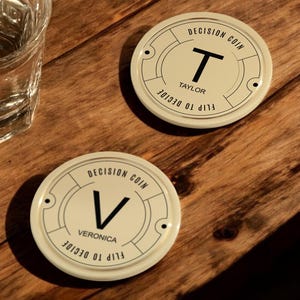 May include: Two round, cream-colored decision coins with black lettering. The top coin reads "Decision Coin" and "Flip to Decide" with the letter "T" in the center and the name "Taylor" below. The bottom coin reads "Decision Coin" and "Flip to Decide" with the letter "V" in the center and the name "Veronica" below.