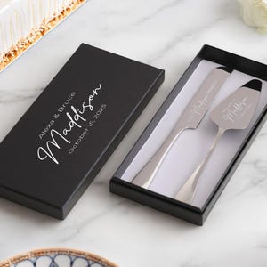 May include: A black box with the text "Alexa & Bruce Madison October 15, 2023" on the top. The box contains two silver cake servers with the text "Alexa & Bruce" and "Mr. & Mrs." engraved on them.
