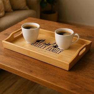 May include: A rectangular bamboo serving tray with two white coffee mugs filled with dark coffee. The tray has handles and a black mountain and text design that reads "BAILEY FAMILY" and "Summit 2023". The tray sits on a wooden table.