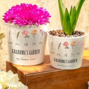 May include: Two white ceramic flower pots with a floral design and the text "Grandma's Garden, joy blooms here". The pots are on a wooden surface. One pot has pink flowers and the other has a green plant.