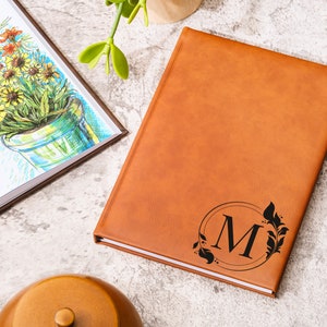 Peut inclure: Un journal en cuir marron avec un monogramme noir de la lettre "M" entour&eacute; d'un motif floral.