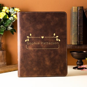 May include: A brown leather portfolio with gold lettering that reads "Sophia Patterson." The portfolio has a decorative gold design above the name. The background includes books, flowers, and a vase.