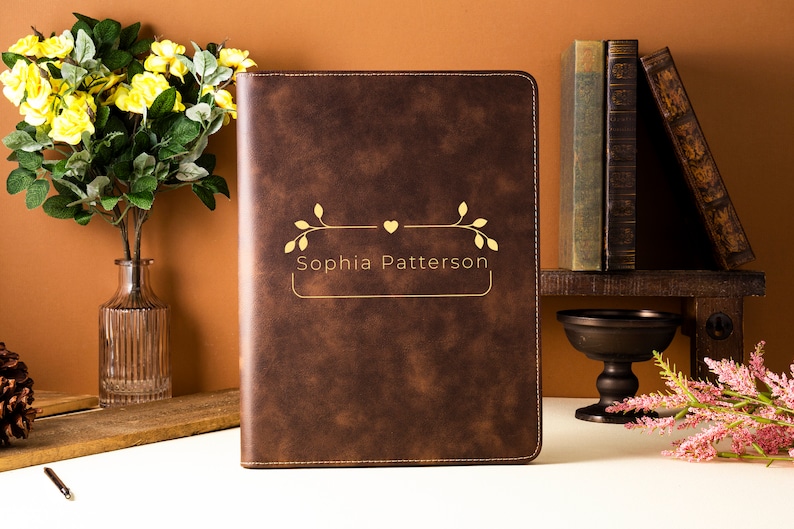 May include: A brown leather portfolio with gold lettering that reads "Sophia Patterson." The portfolio has a decorative gold design above the name. The background includes books, flowers, and a vase.