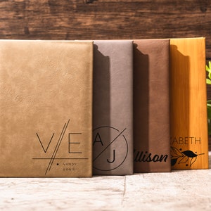 May include: Five personalized leather-bound journals with different initials and names. The journals are arranged from left to right: V/E Vandy Ernie, A/J Allison, Elizabeth, and a wooden journal with a leaf design.