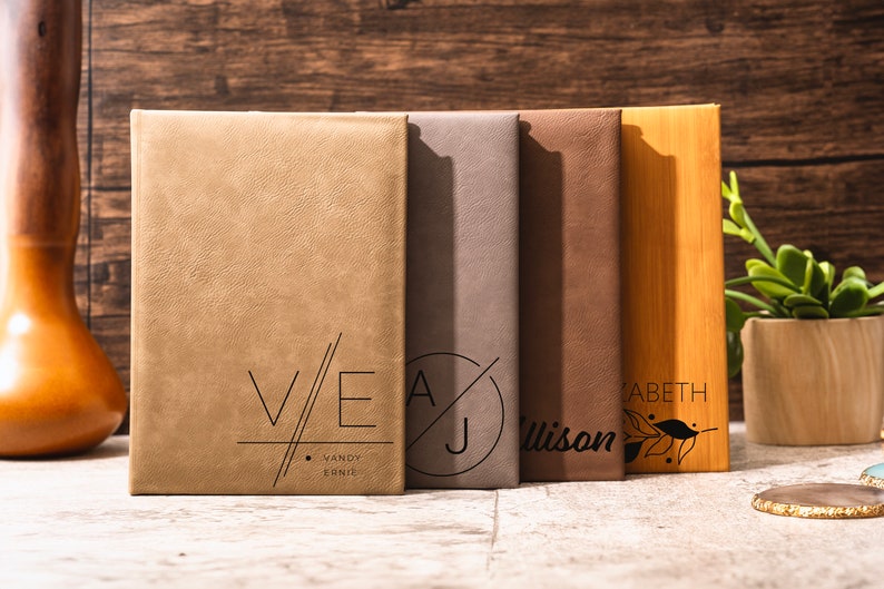 May include: Five personalized leather-bound journals with different initials and names. The journals are arranged from left to right: V/E Vandy Ernie, A/J Allison, Elizabeth, and a wooden journal with a leaf design.