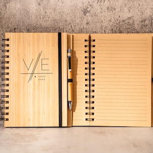 May include: A bamboo notebook with a black spiral binding and a pen attached to the cover. The cover has a black and white graphic design with the initials "V/E" and the text "VANDY ERNIE".