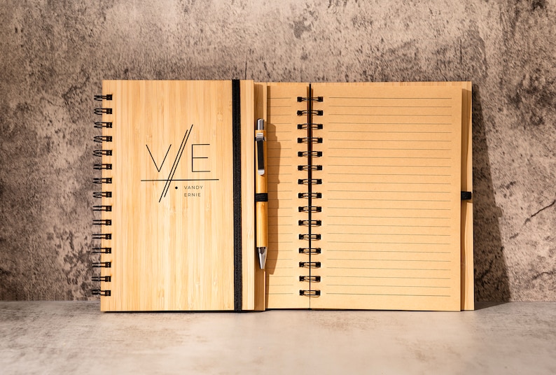 May include: A bamboo notebook with a black spiral binding and a pen attached to the cover. The cover has a black and white graphic design with the initials "V/E" and the text "VANDY ERNIE".