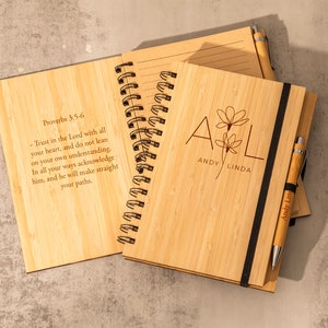 May include: Two bamboo notebooks with a black elastic band and a pen. The front cover of one notebook has the text "Proverbs 3:5-6" and a bible verse. The front cover of the other notebook has the text "A & L Andy Linda" with a floral design.