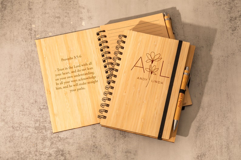 May include: Two bamboo notebooks with a black elastic band and a pen. The front cover of one notebook has the text "Proverbs 3:5-6" and a bible verse. The front cover of the other notebook has the text "A & L Andy Linda" with a floral design.