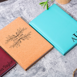 May include: Three personalized notebooks with different covers. One is cork with a floral design and the name "Steve Clark" engraved. Another is a deep red with the name "Alexa Taylor" engraved. The third is a light blue with the initials "AKM" engraved.