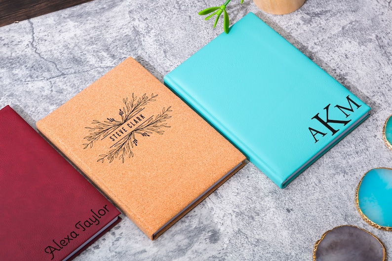May include: Three personalized notebooks with different covers. One is cork with a floral design and the name "Steve Clark" engraved. Another is a deep red with the name "Alexa Taylor" engraved. The third is a light blue with the initials "AKM" engraved.