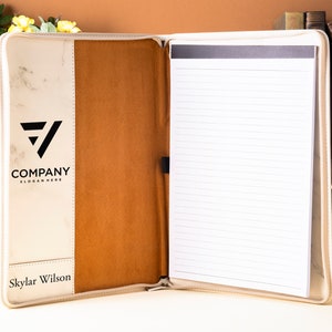 Op de afbeelding: Een witte en bruine leren portfolio met een notitieblok en een pennenhouder. De voorkant heeft een marmerpatroon met een zwart logo met de tekst "COMPANY SLOGAN HERE". De naam "Skylar Wilson" staat onderaan de voorkant gedrukt.