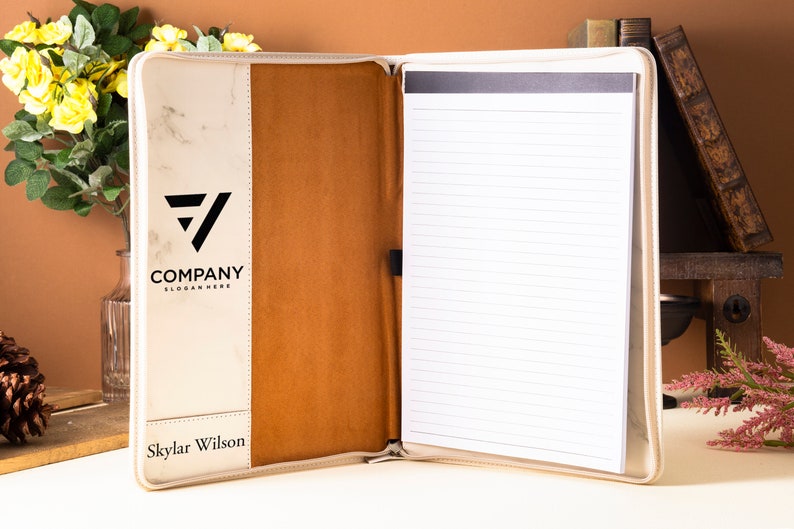 Op de afbeelding: Een witte en bruine leren portfolio met een notitieblok en een pennenhouder. De voorkant heeft een marmerpatroon met een zwart logo met de tekst "COMPANY SLOGAN HERE". De naam "Skylar Wilson" staat onderaan de voorkant gedrukt.