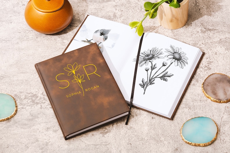 May include: A brown leather journal with the initials "SR" and the names "Sophia Rogan" embossed in gold. The journal is open to reveal a blank page with a black and white drawing of daisies.