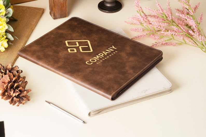 Op de afbeelding: Een bruin lederen notitieboek met een gouden gegraveerd logo met de tekst "COMPANY SLOGAN HERE". Het notitieboek is gesloten en ligt op een wit oppervlak.