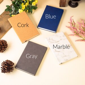 May include: Four different notebooks with various covers: cork, gray, marble, and blue. The notebooks are arranged on a light beige surface.