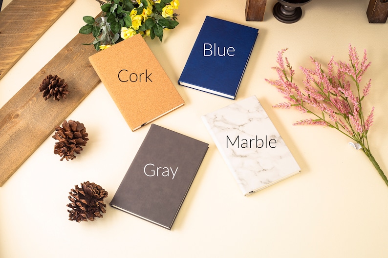 May include: Four different notebooks with various covers: cork, gray, marble, and blue. The notebooks are arranged on a light beige surface.