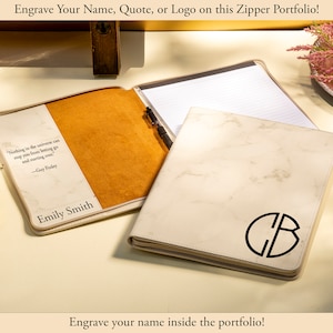 May include: Cream-colored zipper portfolio with a marble-like pattern and a black monogram. The open portfolio reveals a notepad and pen. The interior features a quote and name. Text on the image reads: "Engrave Your Name, Quote, or Logo on this Zipper Portfolio!" and "Engrave your name inside the portfolio!"